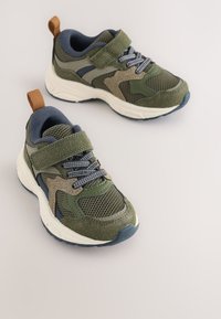 Žalios vaikų sportbačiai su tinklinės ir zomšos tekstūra, turintys velcro dirželį, pilkas detales ir kilpelę ant kulno.