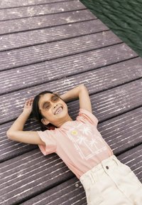 T-shirt rose clair avec un motif de palmier, associée à un short beige. Grandes lunettes de soleil hexagonales en ambre. Quai en bois texturé en arrière-plan.