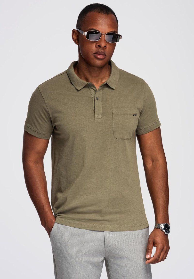 Polo shirt verde oliva con colletto, maniche corte e taschino sul petto; indossato con pantaloni grigio chiaro e occhiali da sole scuri.