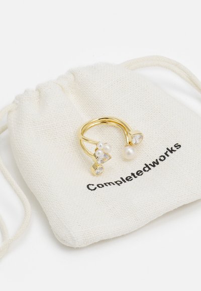 Gold ring featuring three accents: two clear crystals and a white pearl. Presented on a soft white drawstring pouch labeled "Completedworks."