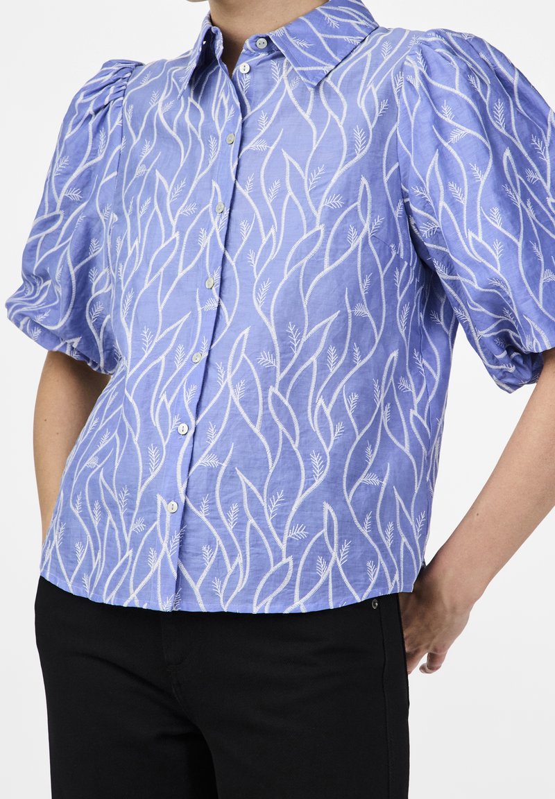 Femme portant un chemisier bleu clair à boutons avec des motifs abstraits de vigne blancs et des manches courtes bouffantes, associé à un pantalon noir.