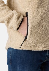Fleece pullover in beige, featuring a textured surface, black zippers, and side pockets. Cuffs are elasticized for a snug fit.