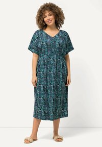 Maxikleid mit einem blauen und grünen Musterdesign, kurzen Ärmeln, V-Ausschnitt und einem elastischen Taillenbund für eine optimale Passform. Kombiniert mit beigen Sandalen.