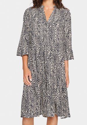 Femme portant une robe mi-longue à imprimé zèbre noir et blanc, manches trois-quarts et col en V, debout devant un fond blanc.