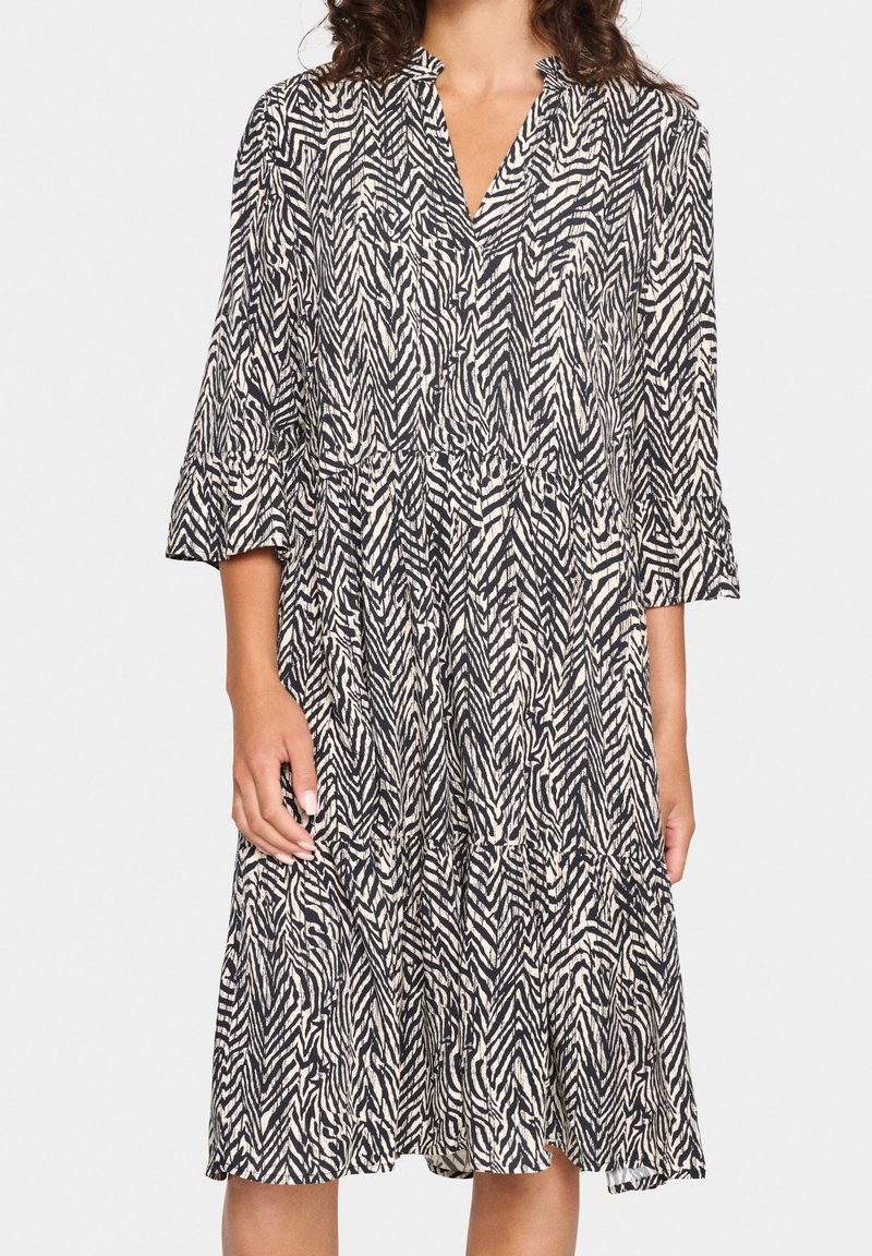 Femme portant une robe mi-longue à imprimé zèbre noir et blanc, manches trois-quarts et col en V, debout devant un fond blanc.