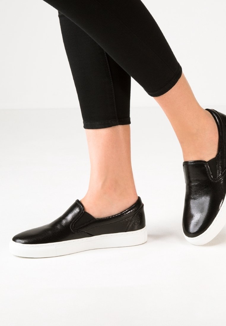 Chaussures noires à enfiler avec une texture brillante et un motif en relief subtil, dotées d'une semelle en caoutchouc blanche et d'une pointe arrondie.