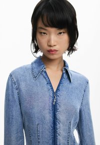 Vestido de mezclilla con cuello de camisa, cierre de cremallera y sutiles detalles deshilachados. Color azul claro con costuras de acento y bolsillos laterales.
