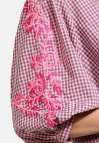 Blusa in fantasia a quadri galletto color malva con ricami floreali rosa sulle maniche. Presenta dettagli arricciati e polsini elasticizzati per una vestibilità ottimale.
