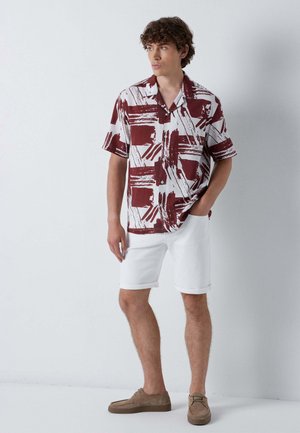 Giovane uomo in piedi con una mano in tasca che indossa una camicia a maniche corte bianca e bordeaux con motivo, pantaloncini bianchi e scarpe marroni.
