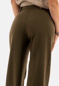 Pantaloni verde oliva con una texture liscia, design sartorial e due tasche posteriori. Dettaglio visibile del bottone in vita.