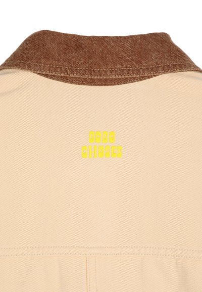 Beige plagg med en brun krage, med gul textur för texten "BOBO CHOSES" på baksidan. Tyget verkar slätt med ett subtilt mönster.