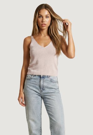 Jonge vrouw met lang blond haar, gekleed in een lichtbeige mouwloze V-hals top en lichtblauwe jeans met hoge taille, poserend tegen een effen achtergrond.