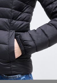 Άτομο που φοράει μαύρο καπιτονέ μπουφάν με το χέρι μέσα στην πλαϊνή τσέπη, σε συνδυασμό με μπλε τζιν.