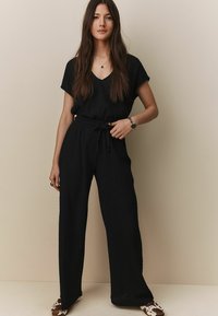 Černý dvoudílný outfit, který se skládá z texturovaného topu s krátkými rukávy a volnými kalhotami s vázáním v pase, doplněný vzorovanými balerínami.