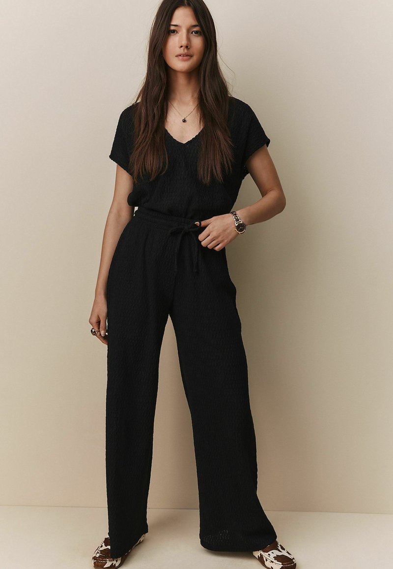 Černý dvoudílný outfit, který se skládá z texturovaného topu s krátkými rukávy a volnými kalhotami s vázáním v pase, doplněný vzorovanými balerínami.