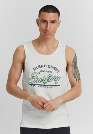 Mann mit kurzen dunklen Haaren, trägt ein weißes Tanktop mit dem Schriftzug "Blend Denim Surfing" und hat ein Tattoo am oberen rechten Arm.