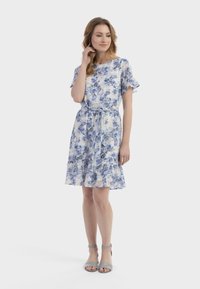 Bloemenjurk met blauwe en witte patronen, korte gerimpelde mouwen, een strakke taille en een uitlopende zoom. Gecombineerd met grijze haksandalen.