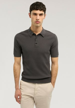 Joven con una camisa polo de manga corta texturizada de color gris oscuro y pantalones beige claro, de pie con una mano en el bolsillo.