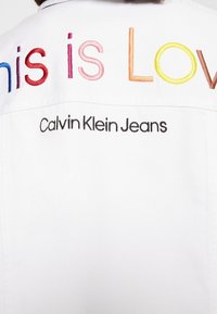 Valkoinen farkkutakki, jossa on monivärisiä brodeerattuja tekstejä "tämä on Rakkautta" ja "Calvin Klein Jeans" mustalla alla.