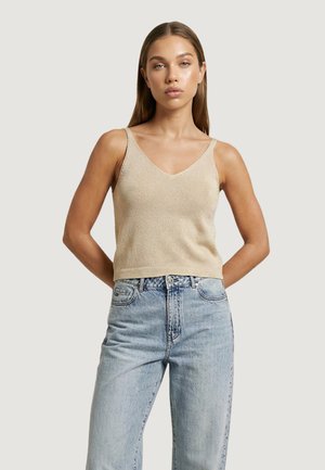 Giovane donna con capelli lisci che indossa un top beige senza maniche a scollo a V e jeans azzurri a vita alta, in piedi con le mani dietro la schiena.