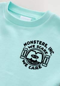 Jasnoniebieska bluza z czarnym haftowanym logo z napisem "MONSTERS, INC. WE SCARE BECAUSE WE CARE" oraz okrągłym wzorem potwora.