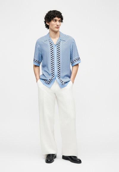 Άνδρας που φοράει ανοιχτό μπλε πουκάμισο με κοντά μανίκια και σχέδια, λευκό παντελόνι με φαρδιά μπατζάκια και μαύρα παπούτσια, στέκεται με τα χέρια στις τσέπες.