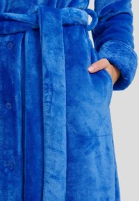 Bata de felpa azul con una textura suave, cierre de botón, cinturón y bolsillos laterales. La tela parece gruesa y acogedora.