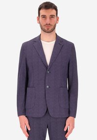 Uomo che indossa un giubbotto di costume blu navy a righe e pantaloni abbinati sopra una t-shirt bianca a girocollo.
