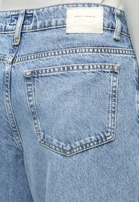 Dettaglio della tasca posteriore di jeans in denim azzurro chiaro con cuciture visibili, passanti per cintura, rivetti e etichetta del marchio sbiadita in vita.