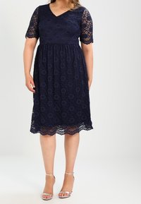 Kobieta ubrana w granatową koronkową sukienkę do kolan z krótkimi rękawami oraz srebrne szpilki, stojąca na białym tle.