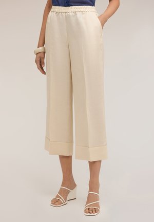 Femme portant un pantalon crème à jambes larges raccourcies avec ourlets retroussés, une blouse bleu marine, des sandales compensées beiges et un bracelet crème au poignet gauche.
