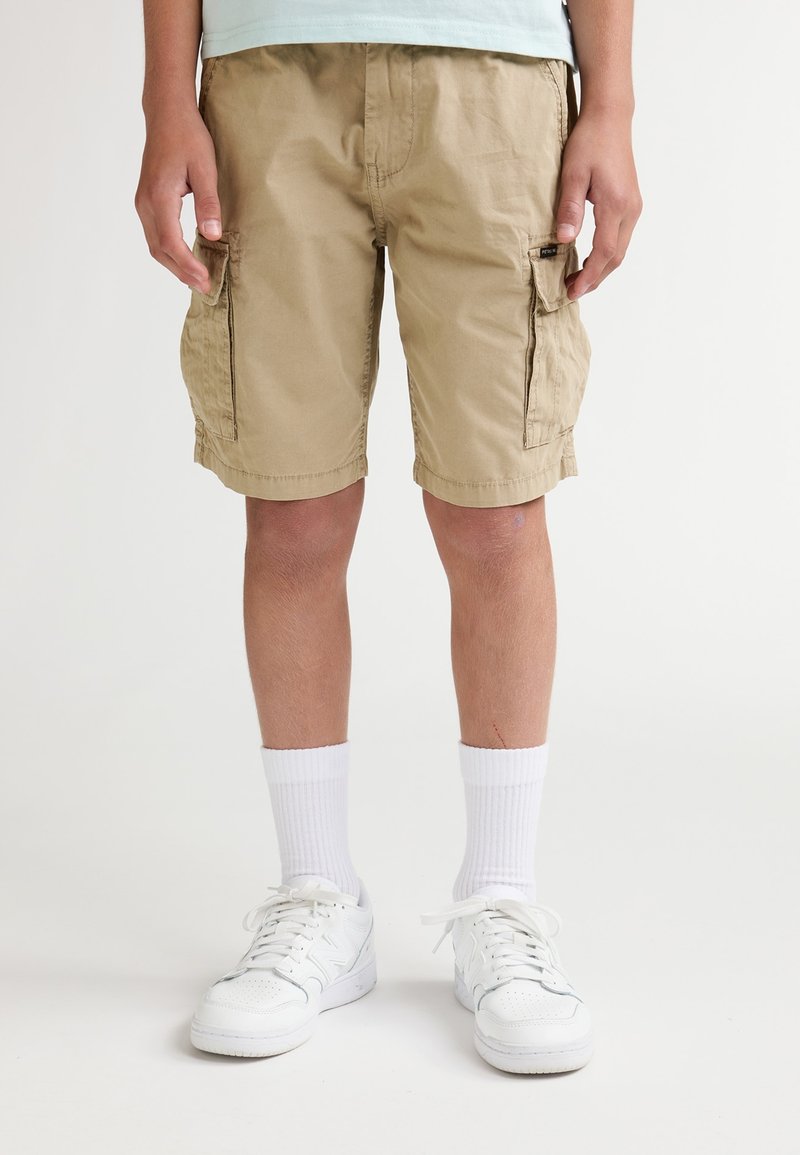 Shorts cargo beiges avec poches latérales, en coton, longueur jusqu'aux genoux. Portés avec des chaussettes blanches montantes et des baskets blanches.