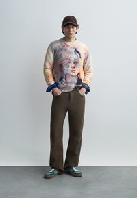 Πλεκτό πουλόβερ με πολύχρωμο πορτρέτο, συνδυασμένο με παντελόνι καφέ σκούρου χρώματος με φαρδιά πόδια και τυρκουάζ αθλητικά παπούτσια με λευκά κορδόνια. Φορά επίσης καφέ καπέλο.