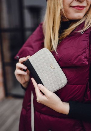 Gray zippered wallet with a textured pattern, featuring a black accent and a gold logo, held in hands against a maroon puffer vest background.