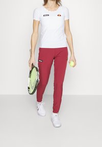 Vit scallop-t-shirt med svart och röd logotyp, matchad med figurnära bourgognebyxor och vita skor, hållande en tennisklubba och boll.