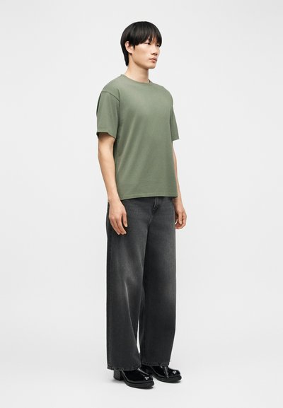 Muž stojaci, oblečený v olivovo zelenej tričku, širokých čiernych vyblednutých džínsoch a čiernych topánkach so štvorcovou špičkou na jednoduchom svetlom pozadí.