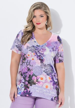 Kvinde med langt blond hår iført en lilla blomstret V-hals skjorte med tekst og lyse lilla bukser, stående mod en hvid baggrund.