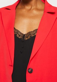 Blazer rouge avec un col cranté, doté d'un seul bouton et d'une texture lisse, superposé sur un haut noir avec des détails en dentelle et des boutons.