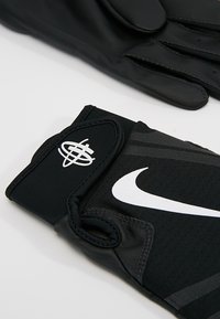 Sorte idrettshansker laget av lær og stoff med teksturert grep, med en hvit Nike-logo og et stripesdesign på håndleddet.