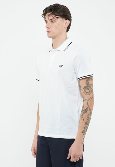 Młody mężczyzna o ciemnych włosach nosi biały polo z czarnym wykończeniem i logo, prezentując szczegółowe tatuaże z wężem i kwiatami na prawej ramieniu.