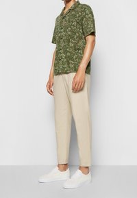 Camicetta verde a maniche corte con un motivo a foglie, abbinata a pantaloni beige a gamba affusolata e sneakers bianche, che mostrano un fit rilassato e un tessuto leggero.