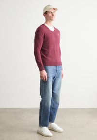 Pulover burgund cu decolteu în formă de V, blugi albastri cu o croială lejeră, adidași albi și o șapcă bej, stând pe un fundal neutru. Modă simplă și casuală.