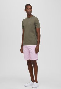 Maglietta di cotone verde oliva con taschino frontale, abbinata a pantaloni corti rosa chiaro e sneakers bianche. Design semplice e casual.