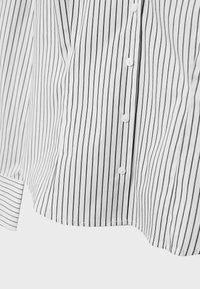 Chemise blanche boutonnée avec de fines rayures verticales noires et des manches longues, montrée de la taille à l'ourlet.
