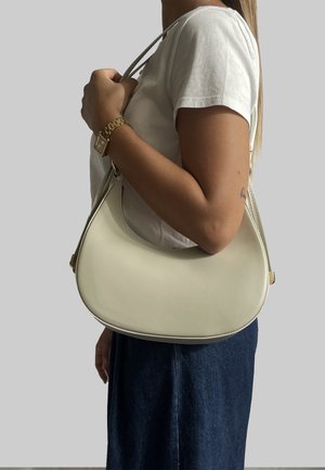 Kremowa skórzana torba na ramię o zakrzywionym kształcie, z pozłacanym okuciem i smukłym paskiem, trzymana przez osobę w białej koszuli.