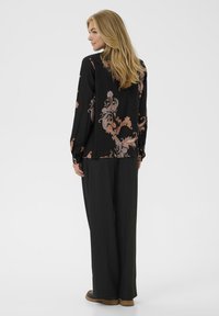 Zwarte top met lange mouwen met een bloemen-paisley patroon in perzik- en lavendeltinten, gecombineerd met losse zwarte broek en bruine schoenen.