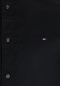 Zwarte overhemdbloes met een gladde textuur, klassieke pasvorm, grijze knopen en een klein rood, wit en blauw logo op de linkerborst.