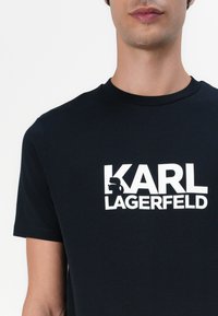Svart bomull t-skjorte med fet hvit "KARL LAGERFELD" tekstlogo, med grafisk silhuettakcent. Klassisk rund hals, kortermet.
