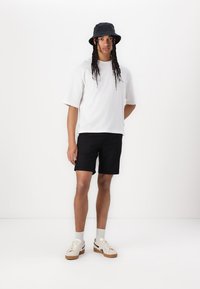 White short-sleeve t-shirt with a relaxed fit, paired with black shorts. Model wears a black bucket hat and beige sneakers with brown soles.