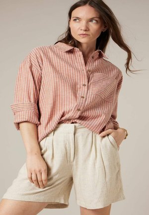 Femme portant une chemise ample à rayures roses et blanches avec manches retroussées et un short beige taille haute, posant avec une main dans la poche.